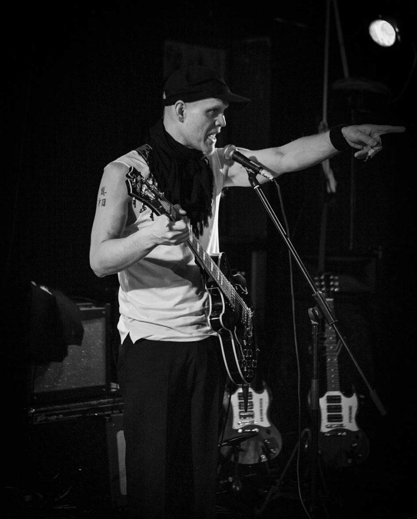 Kuvassa artisti SPK Toivonen lavalla, soittamassa kitaraa ja laulamassa, osoittaen yleisöä mustavalkoisena