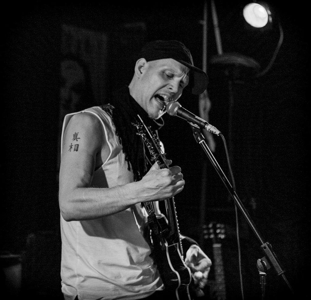 Kuvassa artisti SPK Toivonen lavalla, soittamassa kitaraa ja laulamassa mustavalkoisena
